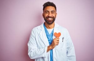 Young indian doctor man holding paper heart standing over isolated pink background with a happy face standing and smiling with a confident smile showing teeth
